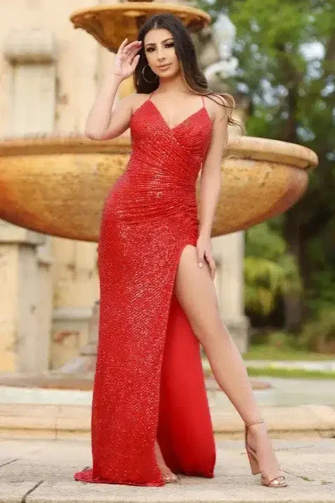 A woman in a sparkling red dress poses confidently, showcasing a high slit while standing near a decorative fountain amidst lush greenery and a stone backdrop.