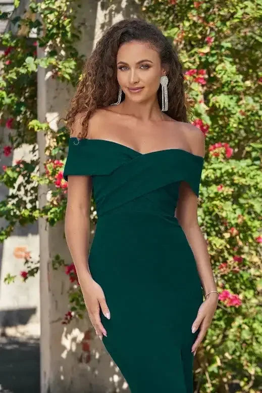 A woman in a deep green off-shoulder gown poses confidently, with long, curly hair and statement earrings. She stands against a backdrop of flowering vines and a textured wall.