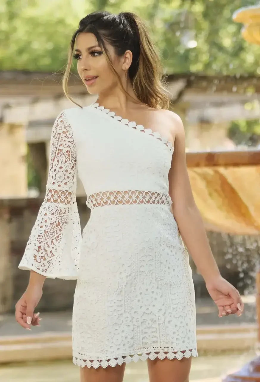 A woman wearing a white lace dress with one shoulder is standing outdoors, smiling confidently. Water flows from a fountain behind her, enhancing the bright, sunny ambiance.
