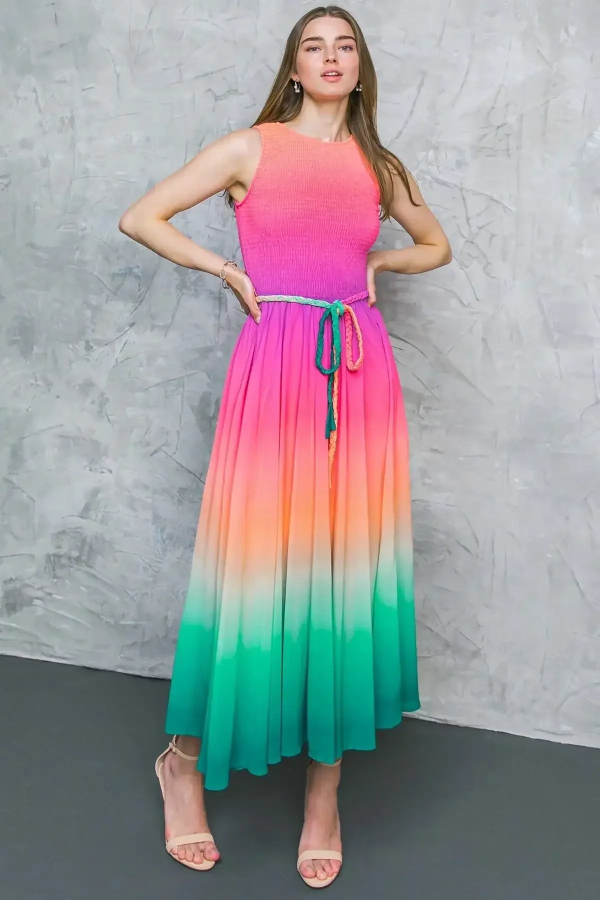 A woman poses confidently in a colorful, gradient dress transitioning from pink to green, highlighting her long hair against a textured gray wall, with a subtle focus on her stylish sandals.