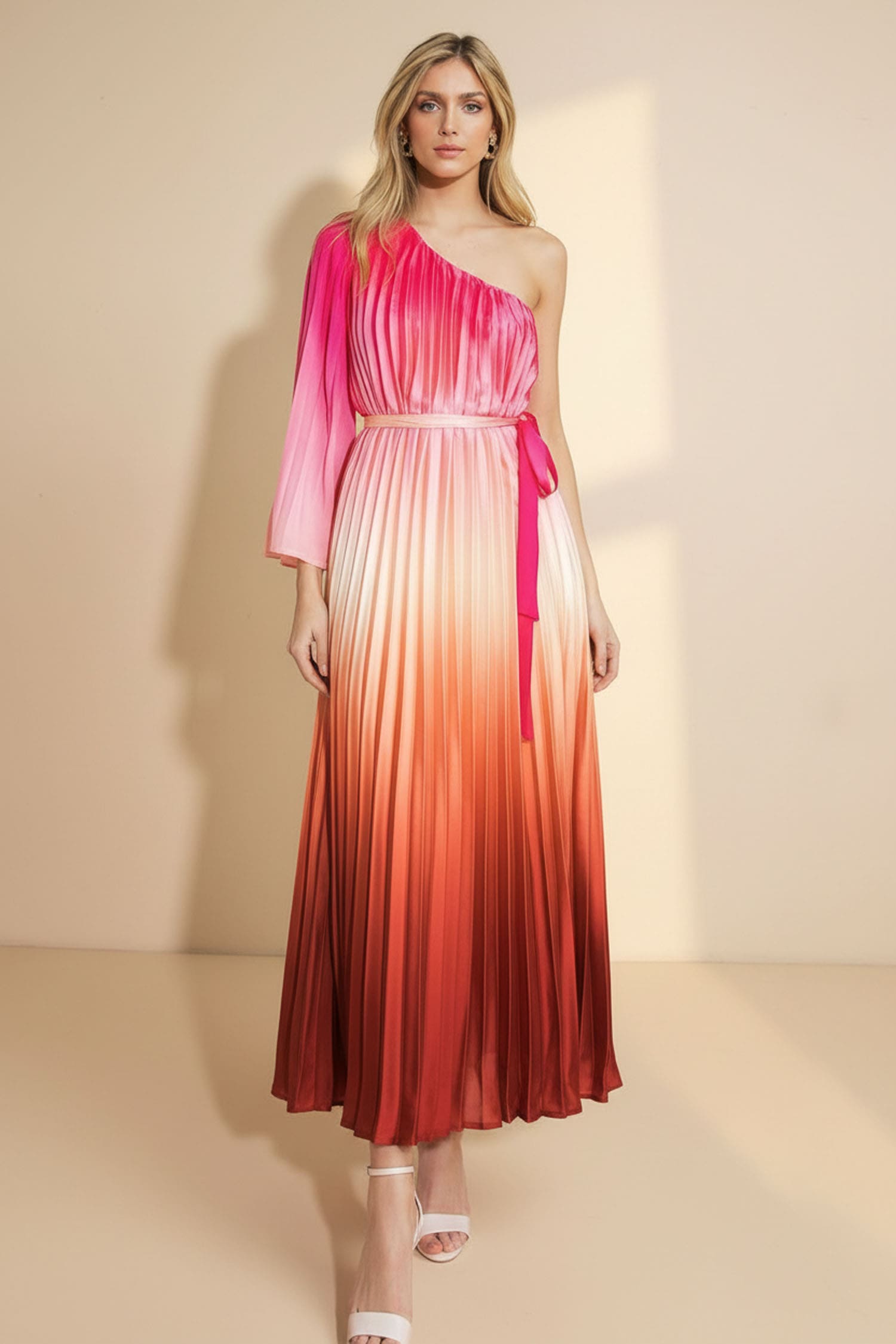 A woman models a one-shoulder pleated maxi dress with a vibrant pink-to-red gradient ombre effect, featuring a matching waist tie, set against a warm beige studio backdrop.