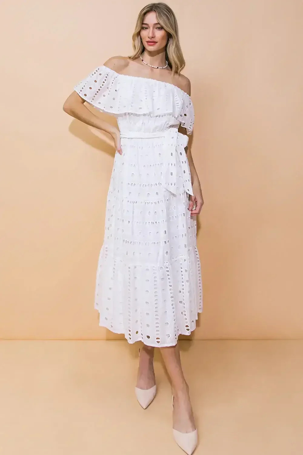 A woman models a white off-the-shoulder dress with ruffled details and lace cutouts. She stands against a soft beige background, one hand on her hip, showcasing an elegant look.