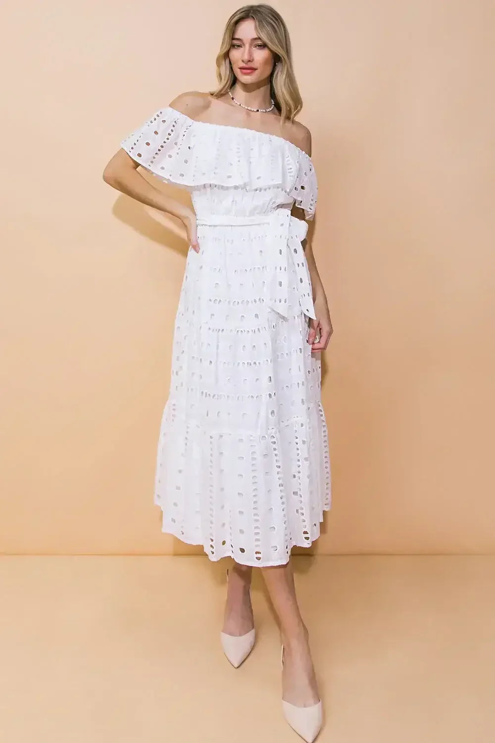 A woman models a white off-the-shoulder dress with ruffled details and lace cutouts. She stands against a soft beige background, one hand on her hip, showcasing an elegant look.