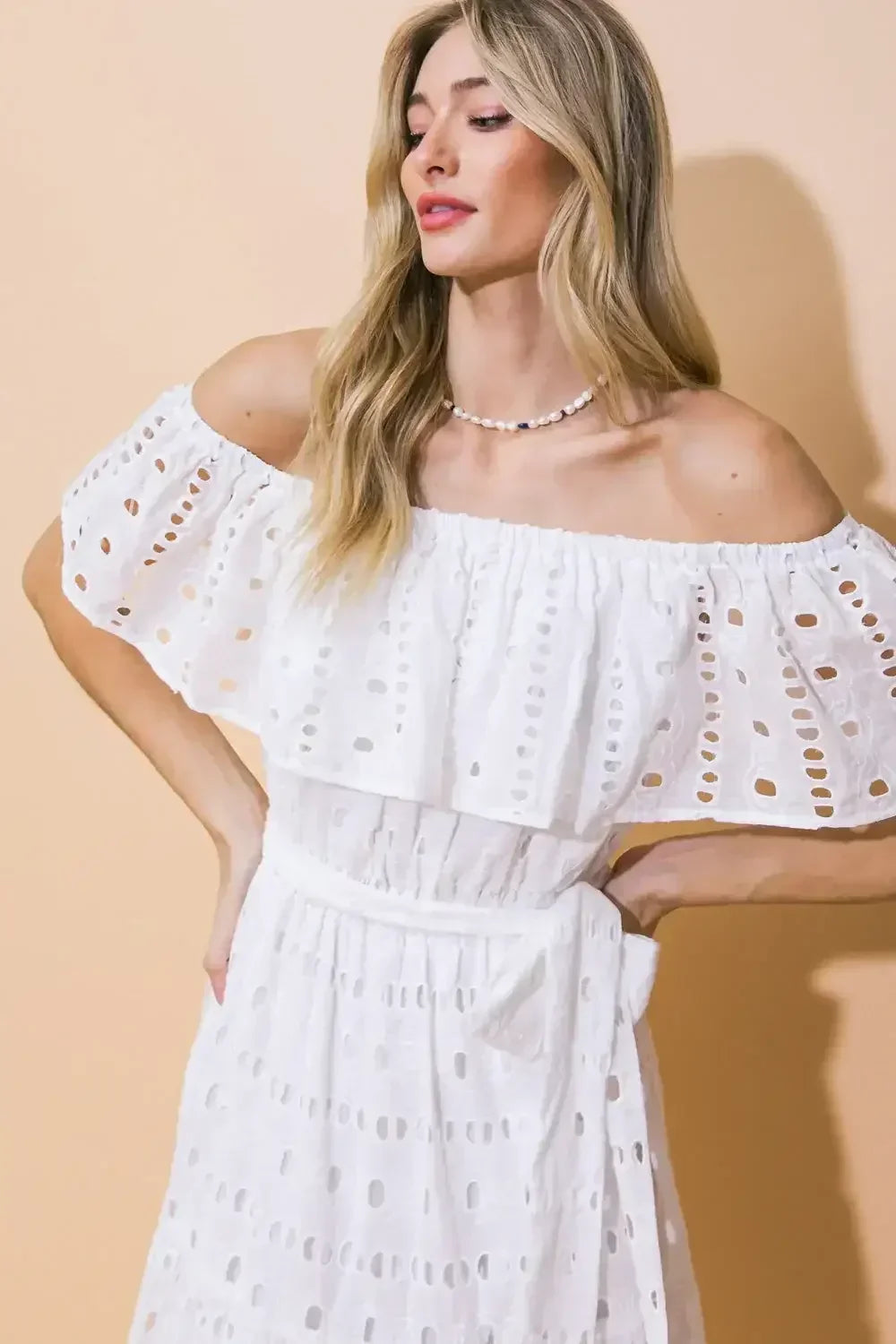 A woman in a stylish white off-the-shoulder dress with eyelet patterns poses confidently. She has long, wavy hair and wears a simple necklace, set against a warm, peach-colored background.
