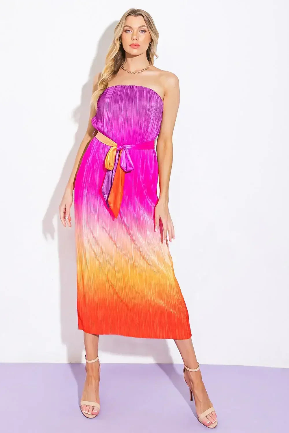 A woman stands confidently in a vibrant, strapless dress that transitions from purple to pink to orange. She pairs it with beige high-heeled sandals, against a plain white backdrop.