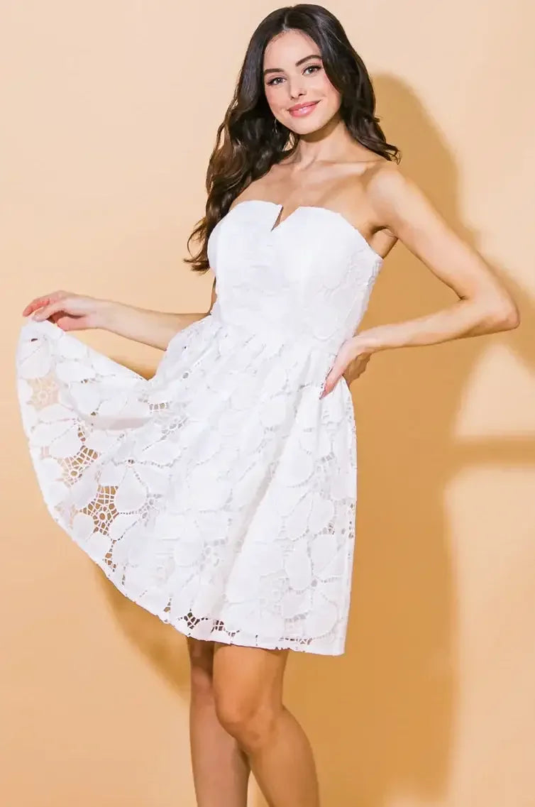 A woman wears a white strapless dress with a floral lace pattern, playfully lifting the hem. She stands against a soft peach background, showcasing her sandals and flowing hair.