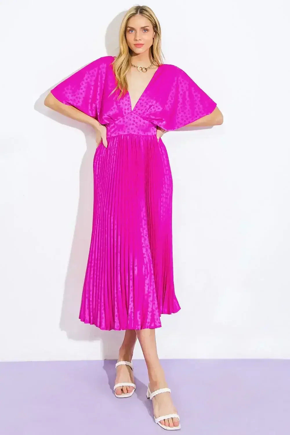 A woman is posing confidently in a vibrant, pleated pink dress with wide sleeves. She stands on a light purple floor against a plain white background, showcasing a stylish, modern look.