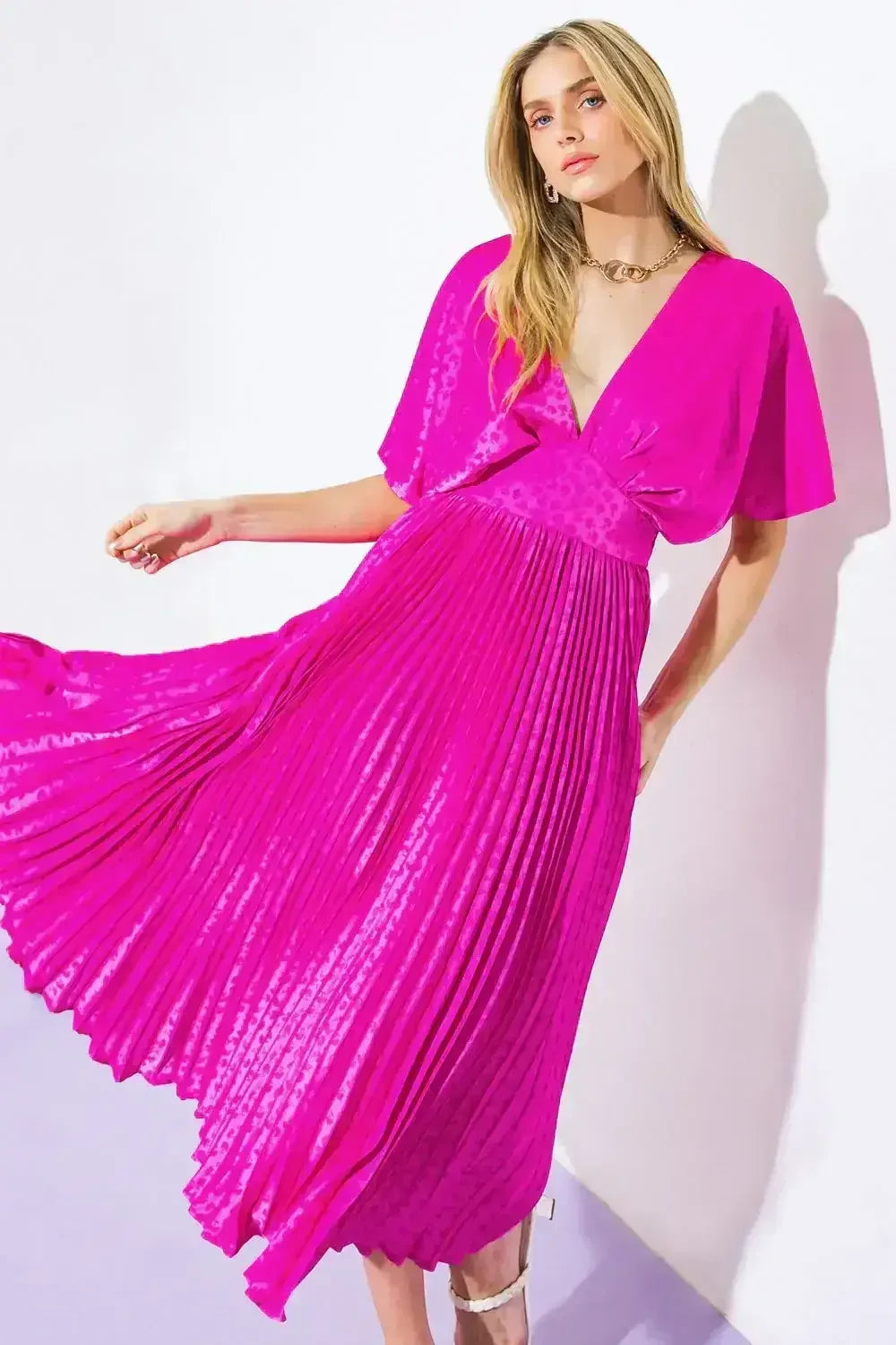 A woman in a vibrant pink, pleated dress twirls gracefully, showcasing the flowing fabric. She stands against a minimalist white wall and light purple floor, enhancing the dress's bold hue.