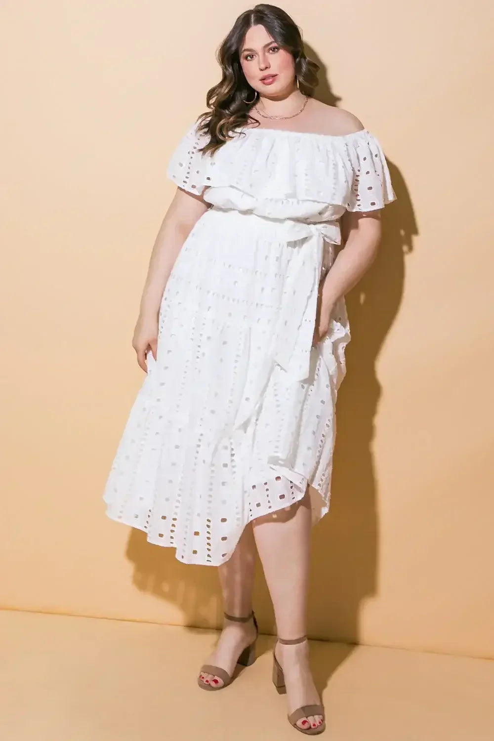 A woman in a white, off-the-shoulder, tiered dress stands confidently, with one hand on her hip, against a soft, beige background. She wears beige block-heeled sandals.