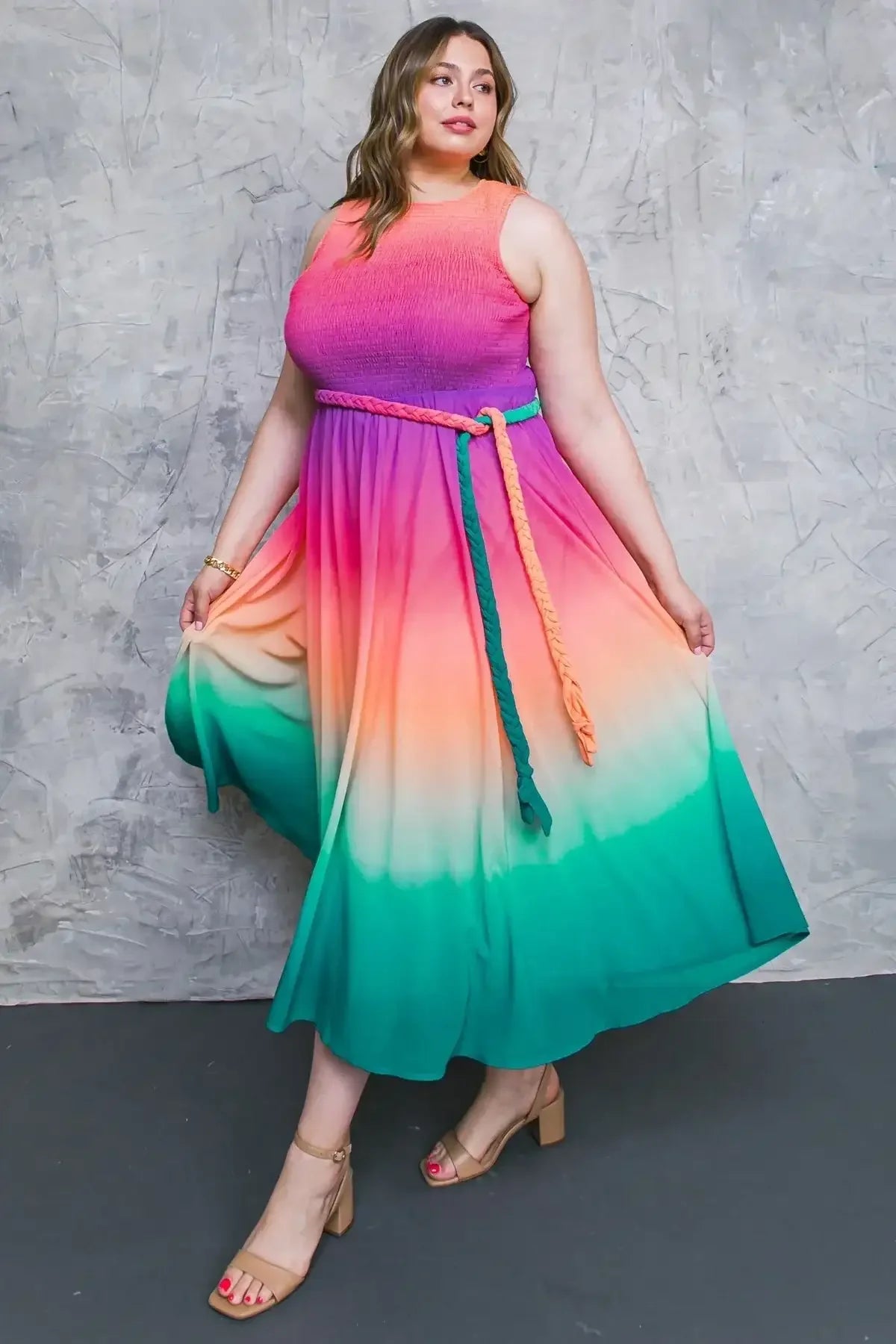 A woman is gracefully posing in a colorful, ombre maxi dress with a fitted bodice that transitions from pink to orange to green. She wears block-heeled sandals and a braided belt. The background features a textured gray wall.