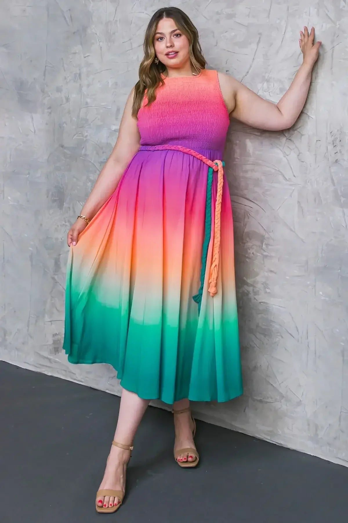 A woman in a colorful gradient dress stands against a textured gray wall, holding the fabric of her dress while wearing strappy sandals, showcasing a stylish look.