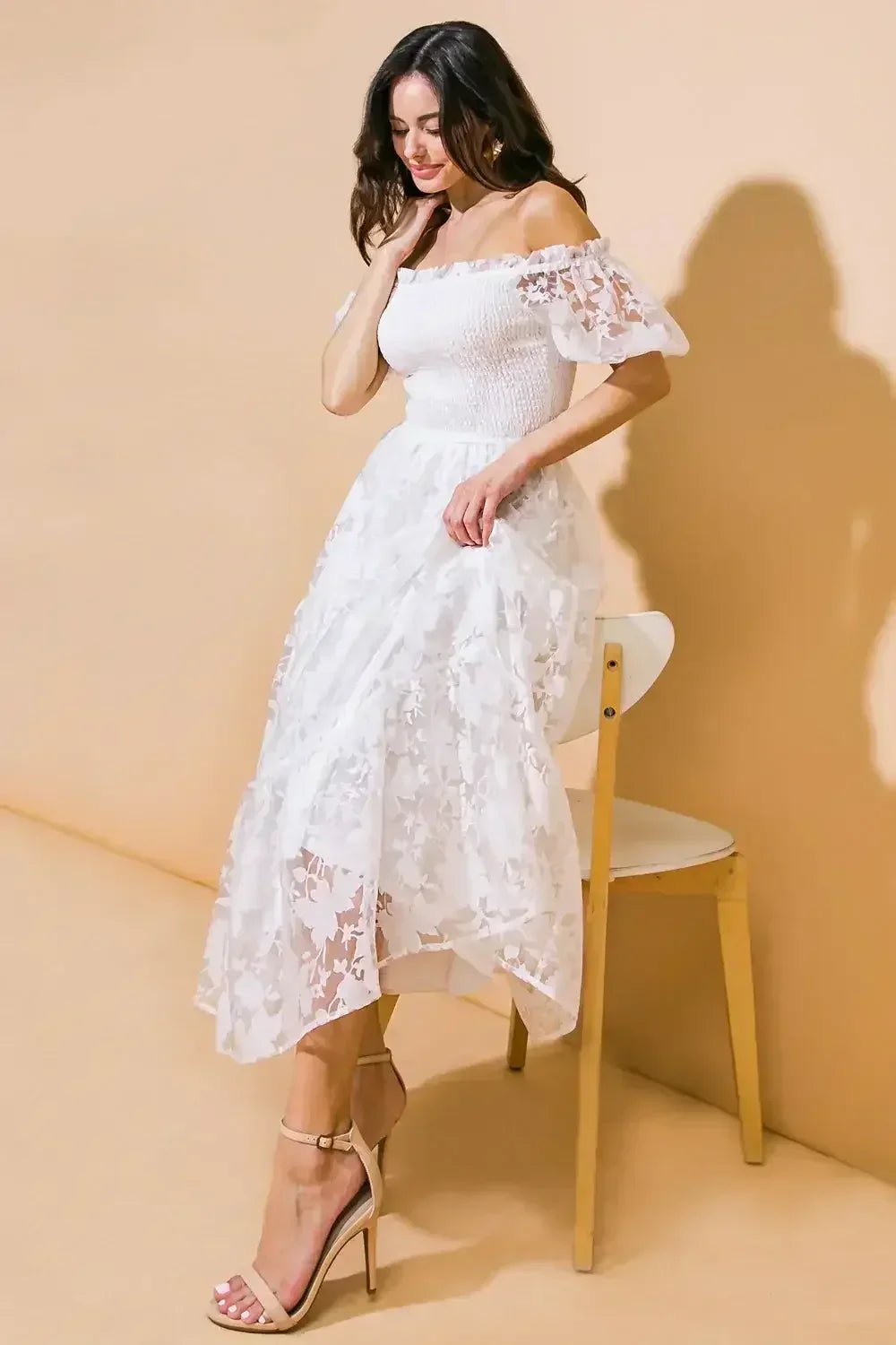 A woman in a white, off-shoulder dress with sheer floral layers is playfully adjusting her hair, standing beside a simple wooden chair against a soft beige background.