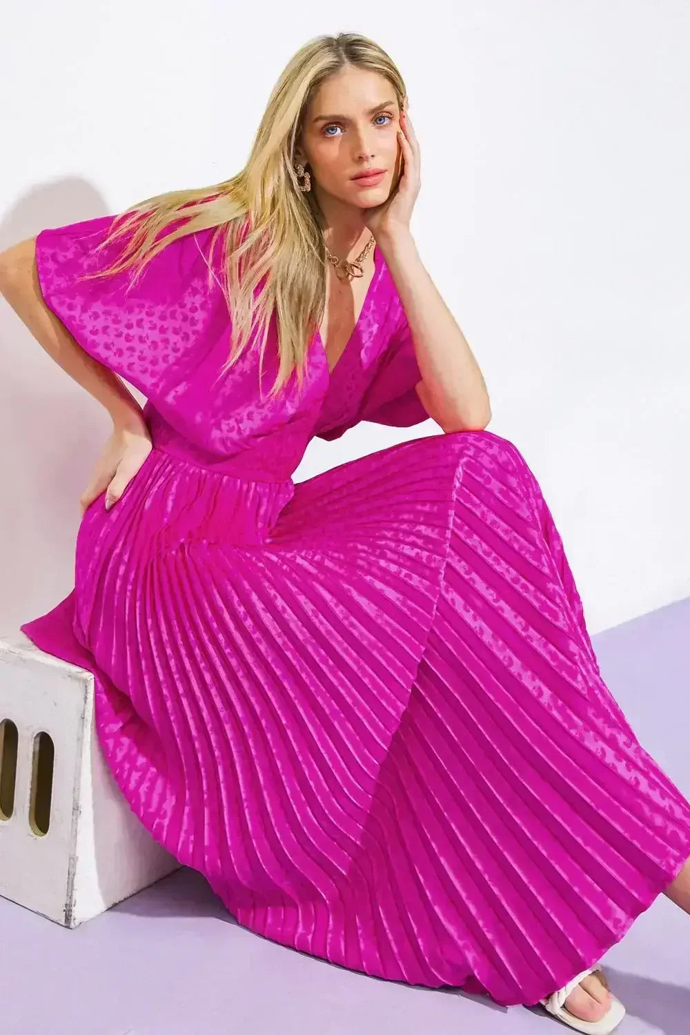 A woman sits elegantly on a light-colored stool, wearing a vibrant pink, pleated maxi dress. She rests her chin on her hand, posing against a minimalistic background.