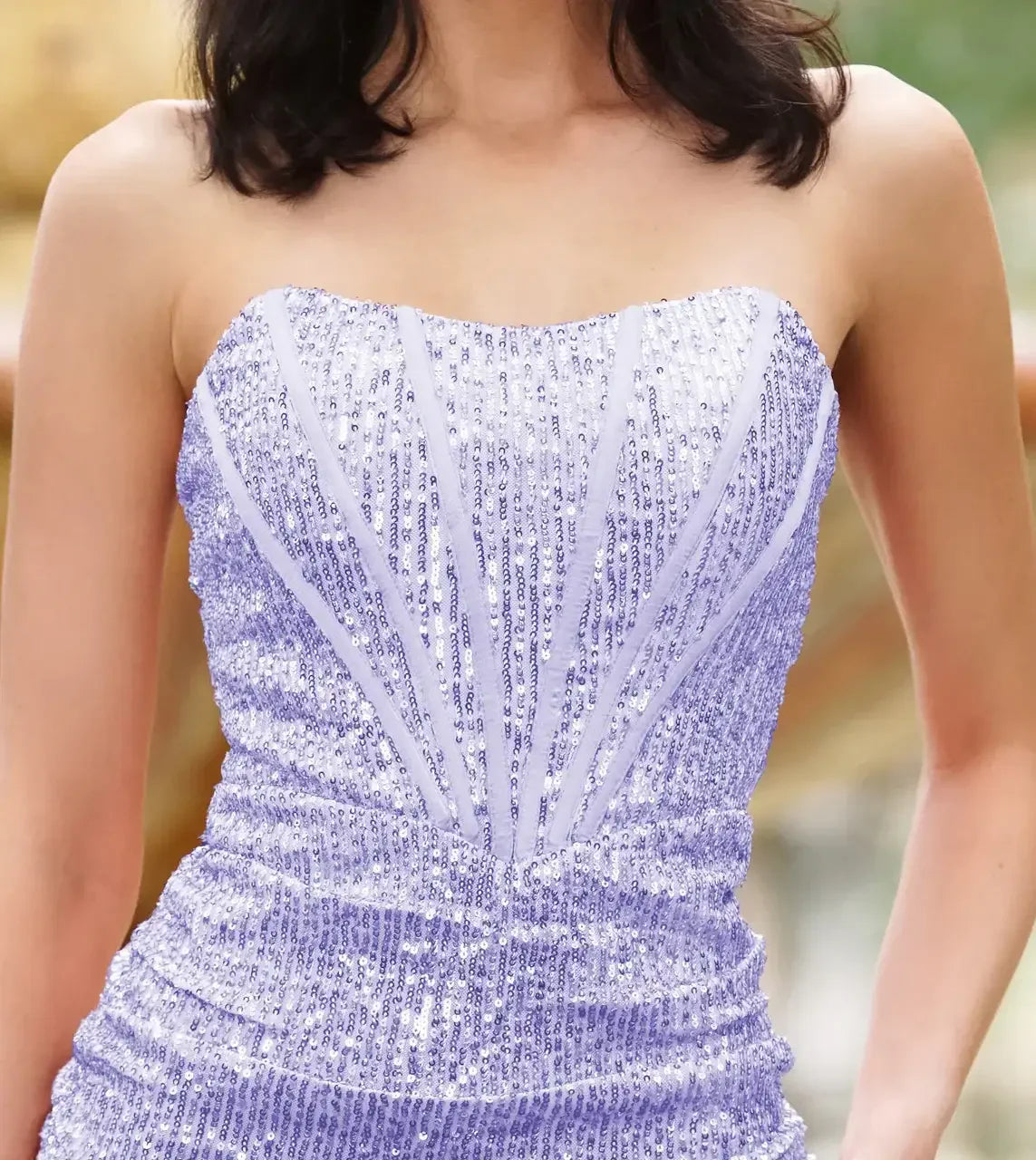 A strapless, sparkly lavender dress with intricate sequins fits closely, showcasing a form-fitting design. The individual is in a blurred outdoor setting, suggesting an elegant atmosphere.