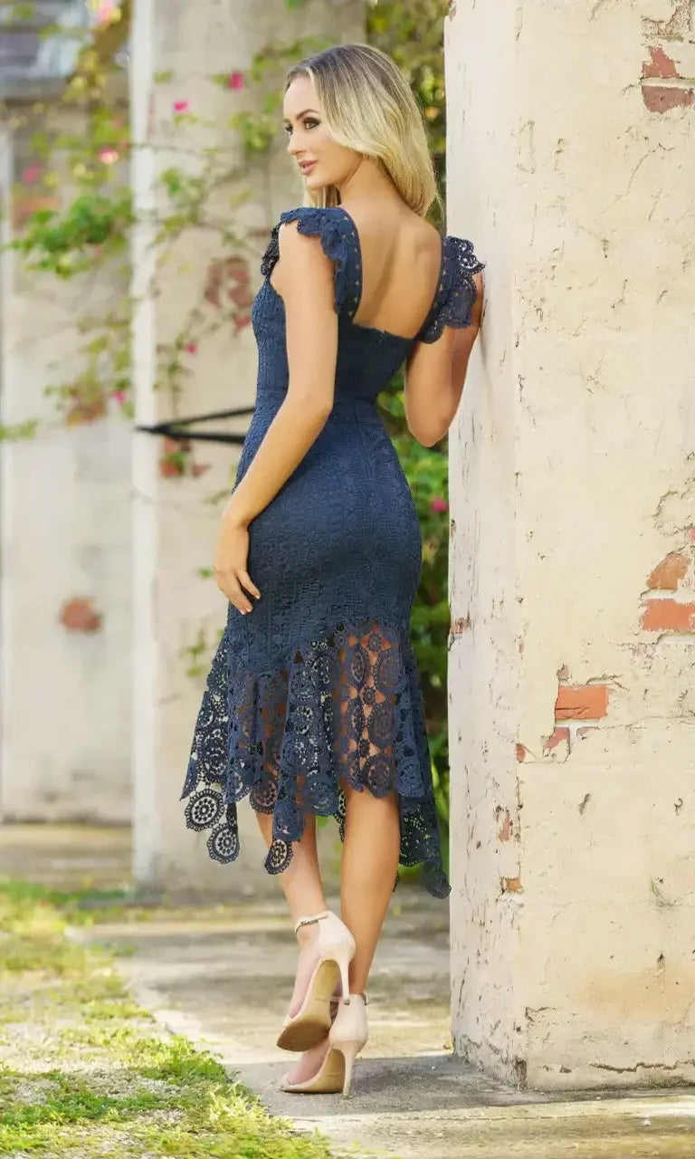 A woman wears a dark lace dress with a flared hem, showcasing a confident pose against a textured wall. The background features soft greenery and rustic elements.