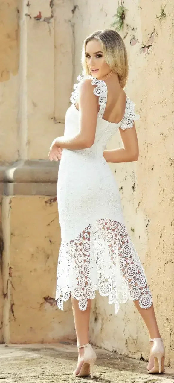 A woman wears a white lace dress with ruffled sleeves, turning to look back over her shoulder. She stands against a textured, muted stone wall.