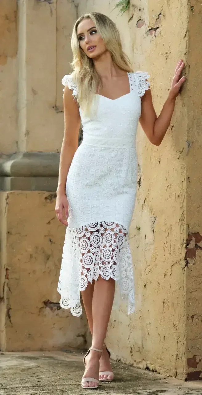 A woman stands against a wall, wearing a white dress with lace detailing. She poses confidently while her long hair cascades over her shoulders. The setting features textured, earthy tones.