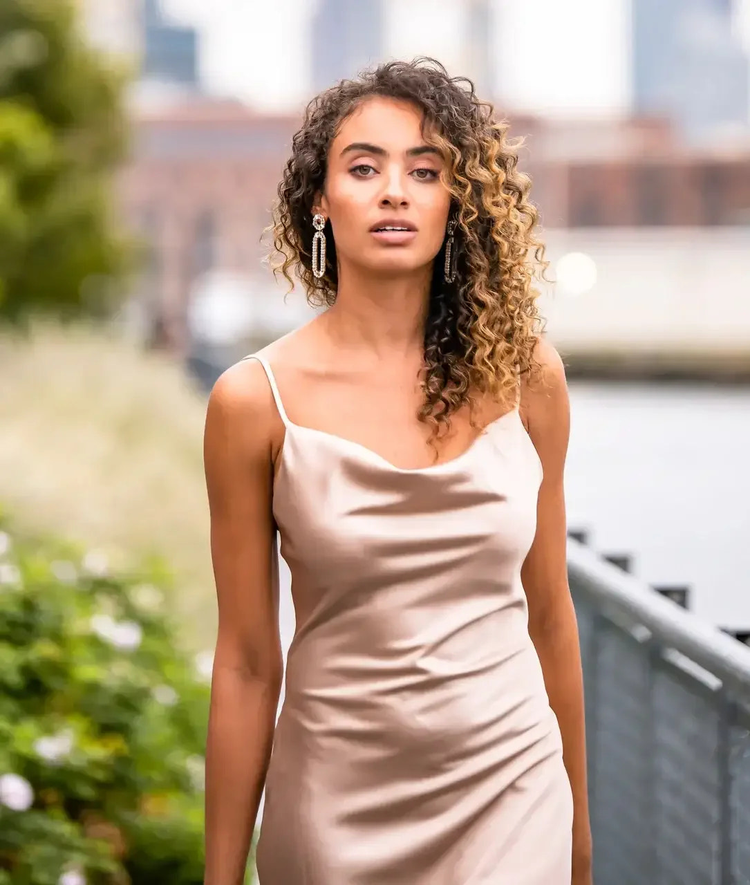 A woman with curly hair walks confidently along a pathway, wearing a sleek satin dress and dangling earrings, with greenery and a waterfront cityscape in the background.