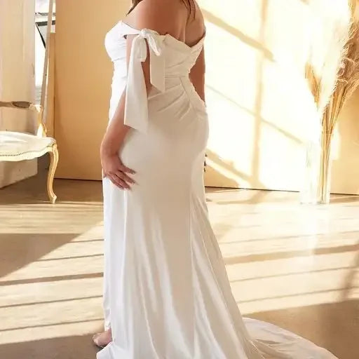 A woman in a white off-the-shoulder gown stands gracefully, displaying a flowing skirt. The scene is illuminated by soft, natural light, with elegant decor in a minimalistic indoor space.