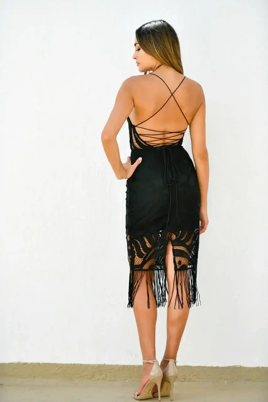 A woman stands with her back to the viewer, wearing a fitted black dress with intricate lace and fringe details. The background features a plain white wall, and she has tan high heels on.