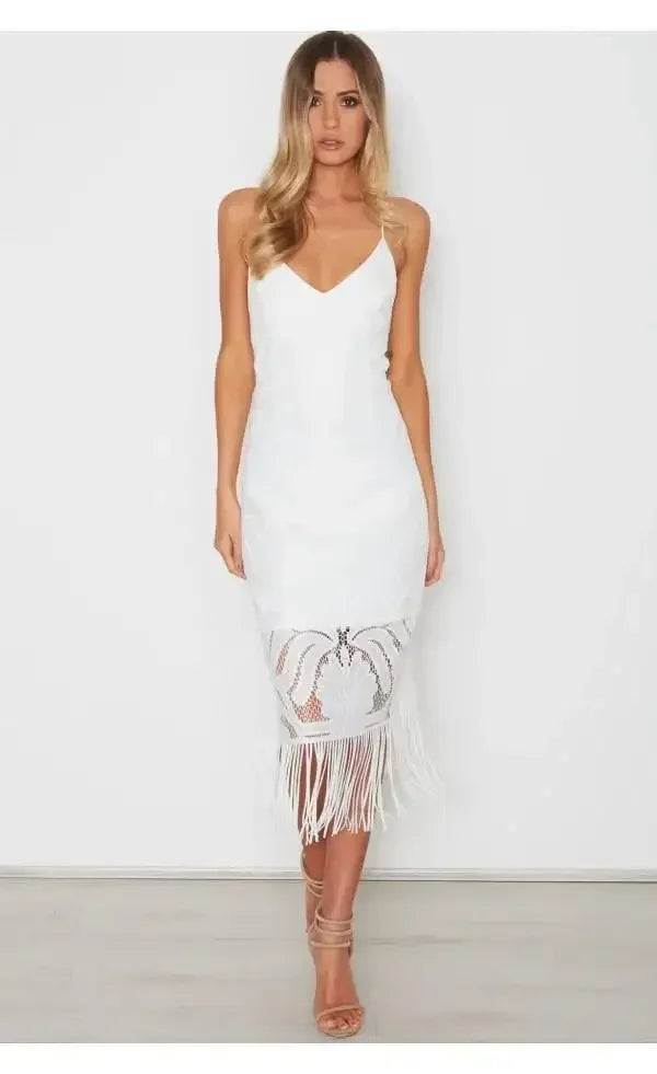 A woman stands confidently in a white dress with a fitted bodice and fringed hem, showcasing a stylish design. She has long, wavy hair and wears strappy heels on a light-colored floor.