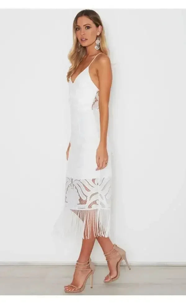 A woman stands in profile, wearing a white, elegant dress with intricate fringe details at the hem. She accessorizes with large earrings and strappy nude heels in a bright, minimalist setting.