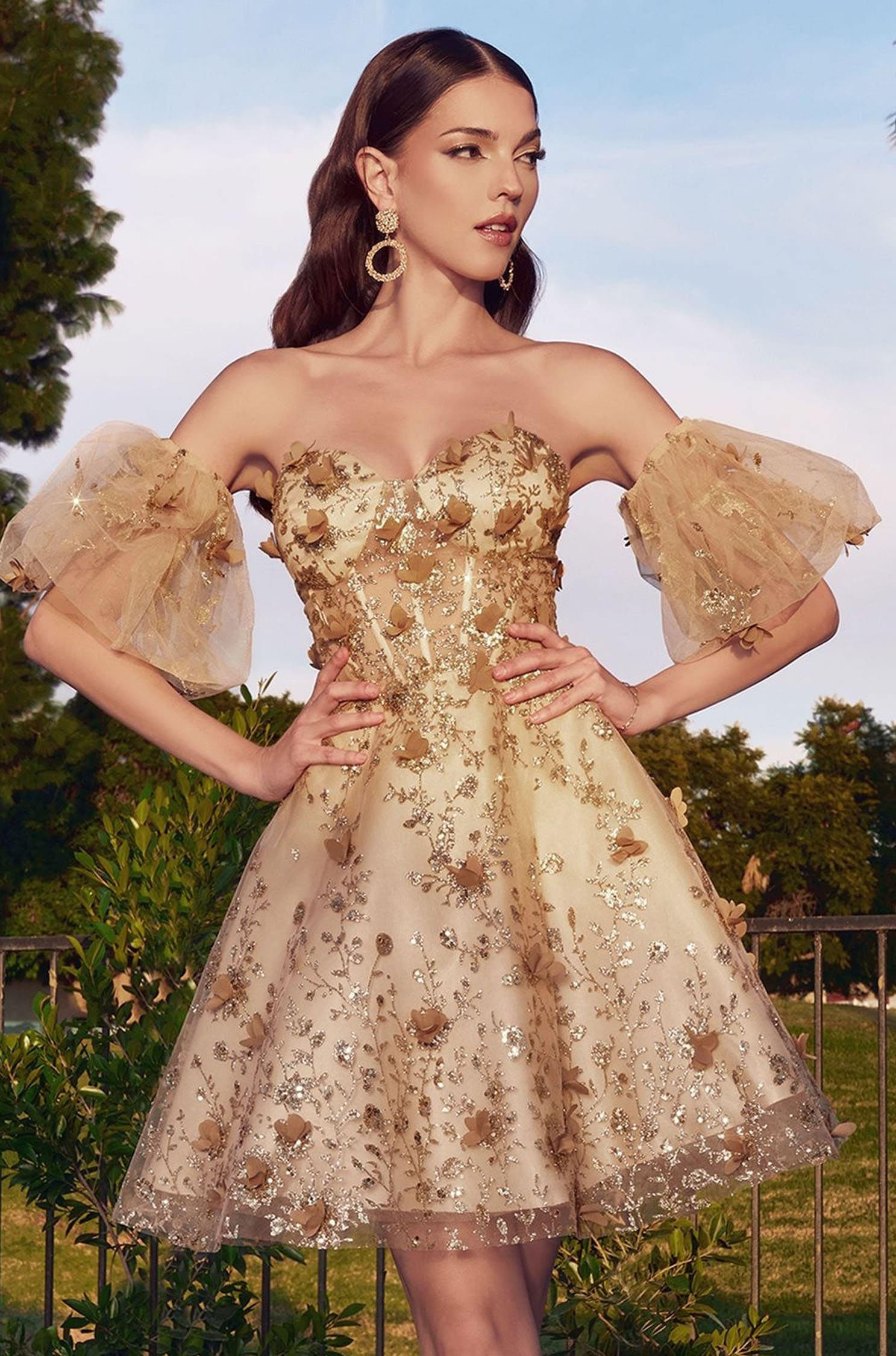 A woman wearing a strapless champagne beige tulle dress with gold floral embroidery and puff sleeves, posing outdoors against a green garden background.