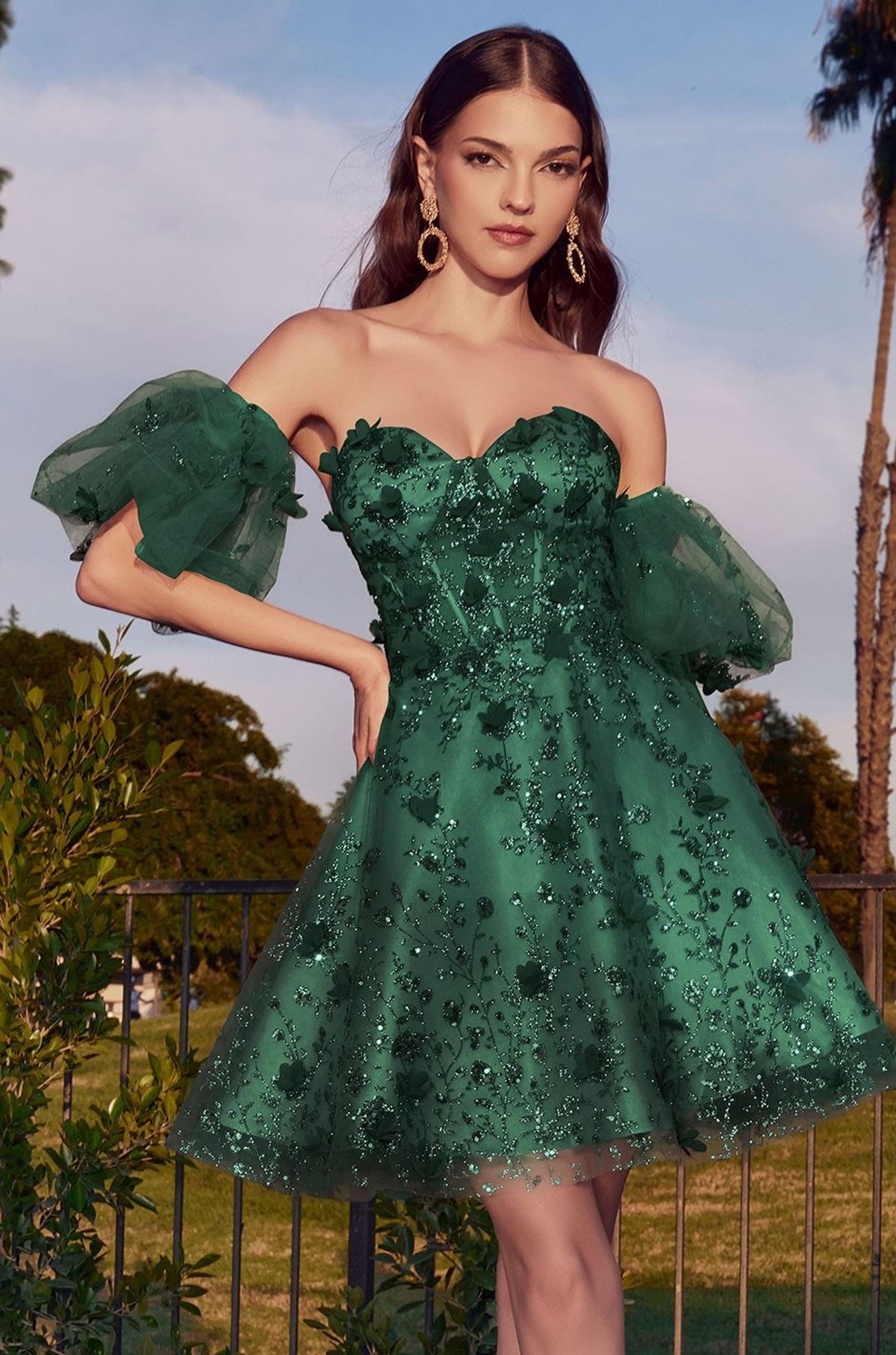 A woman modeling an off-the-shoulder green tulle dress with floral applique and puff sleeves, photographed outdoors against a sky and trees background.