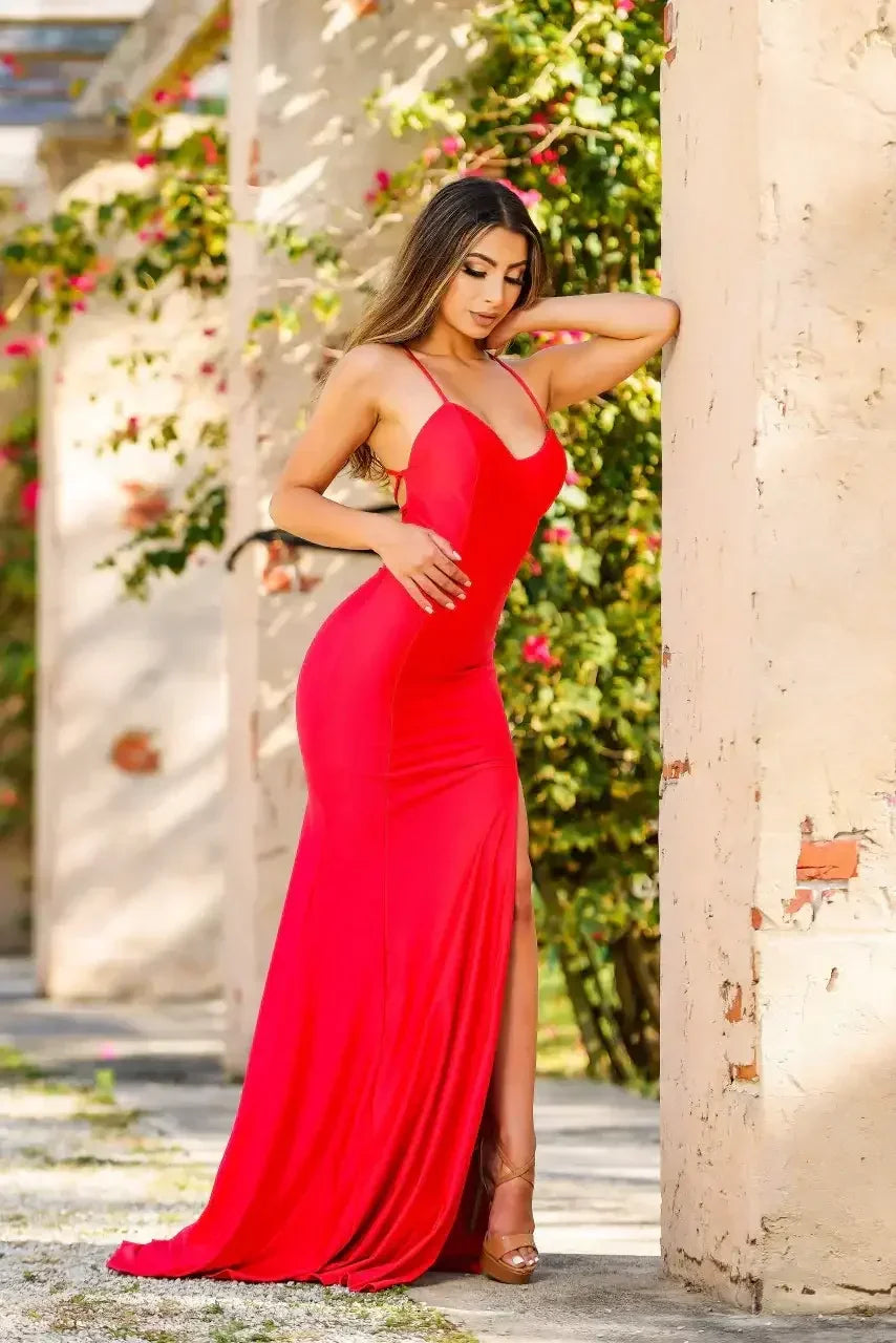A woman in a form-fitting red gown leans against a textured wall, showcasing a confident pose. Lush greenery and pink flowers create a vibrant backdrop in a sunlit outdoor setting.