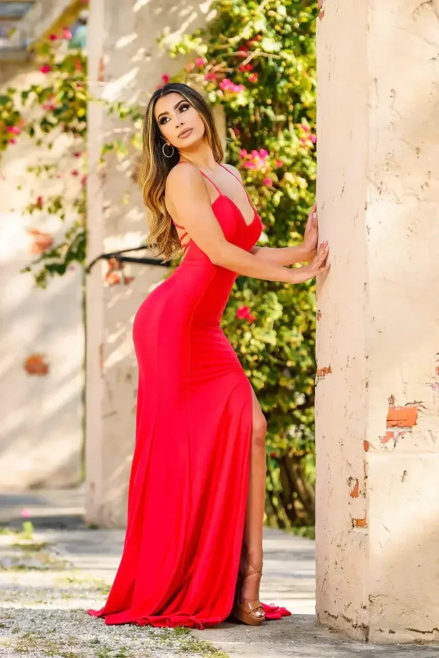 A woman in a fitted red dress poses against a textured wall, with one hand on the wall and the other softly by her side, surrounded by vibrant greenery and pink flowers.