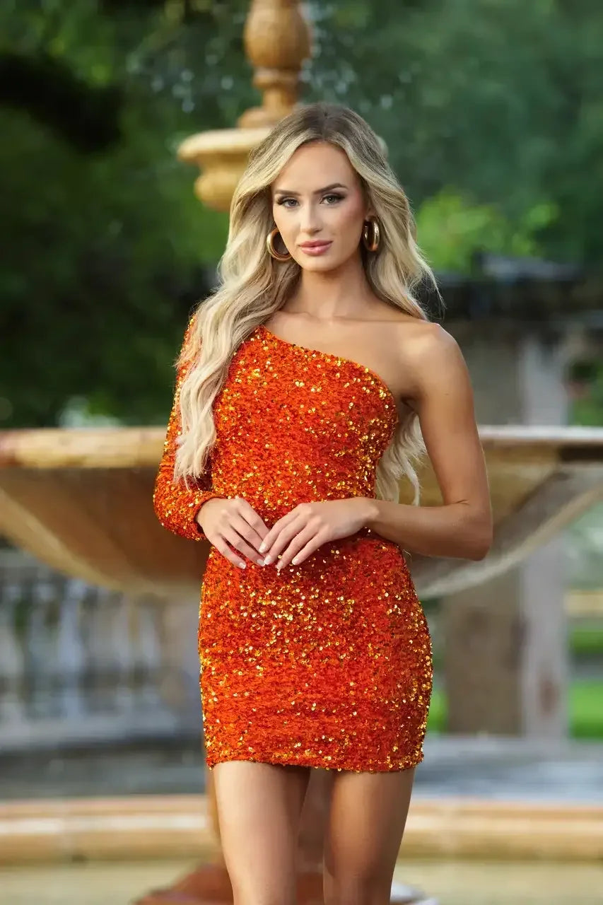 A woman stands confidently in an orange sequined dress, posing with her hands elegantly clasped. She is in a lush garden with a blurred fountain in the background.