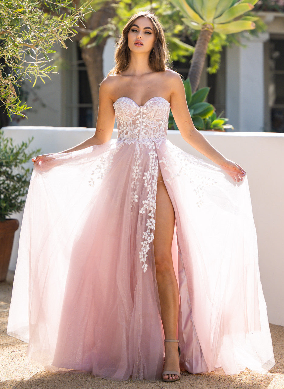 A model wears a strapless, pink gown with floral embroidery, gracefully holding the flowing skirt. The setting features lush greenery and a light-colored backdrop, suggesting an outdoor event.