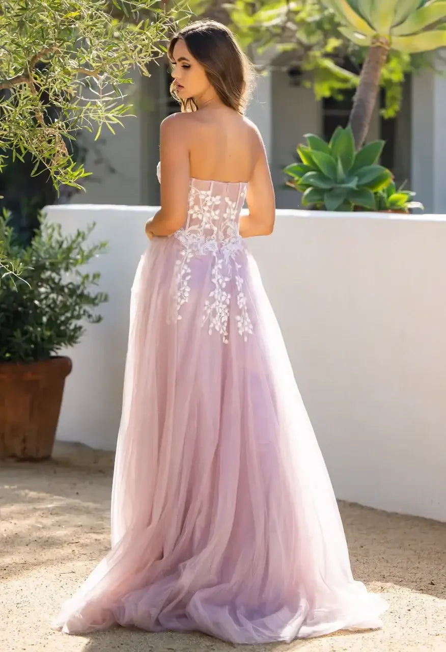 A young woman in a strapless lavender gown stands with her back to the viewer, surrounded by greenery and a smooth white wall, in a sunlit outdoor setting.