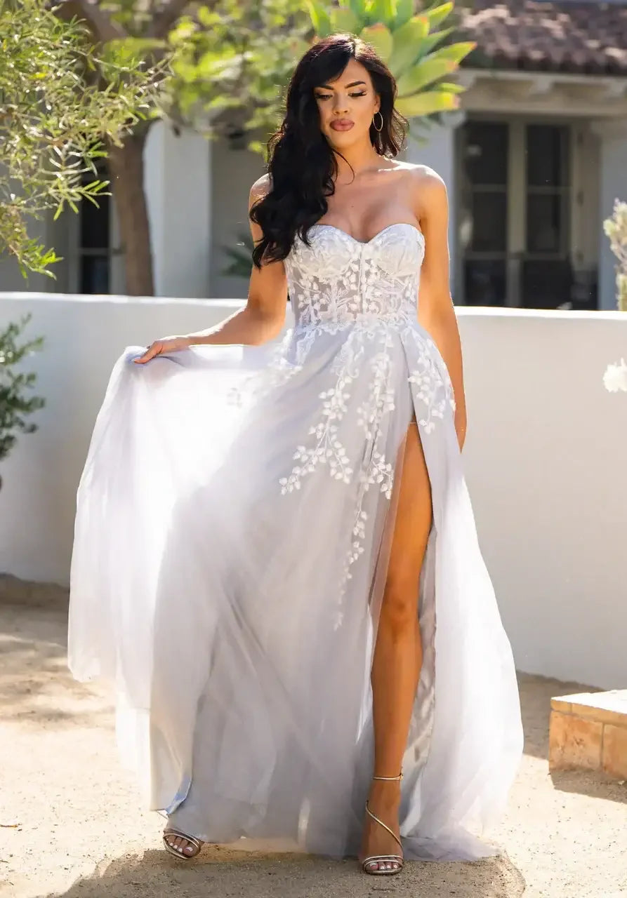 A woman in a flowing, light gray gown stands gracefully, lifting the fabric with one hand. Sunlight filters through surrounding greenery, highlighting her pose in a serene outdoor setting.