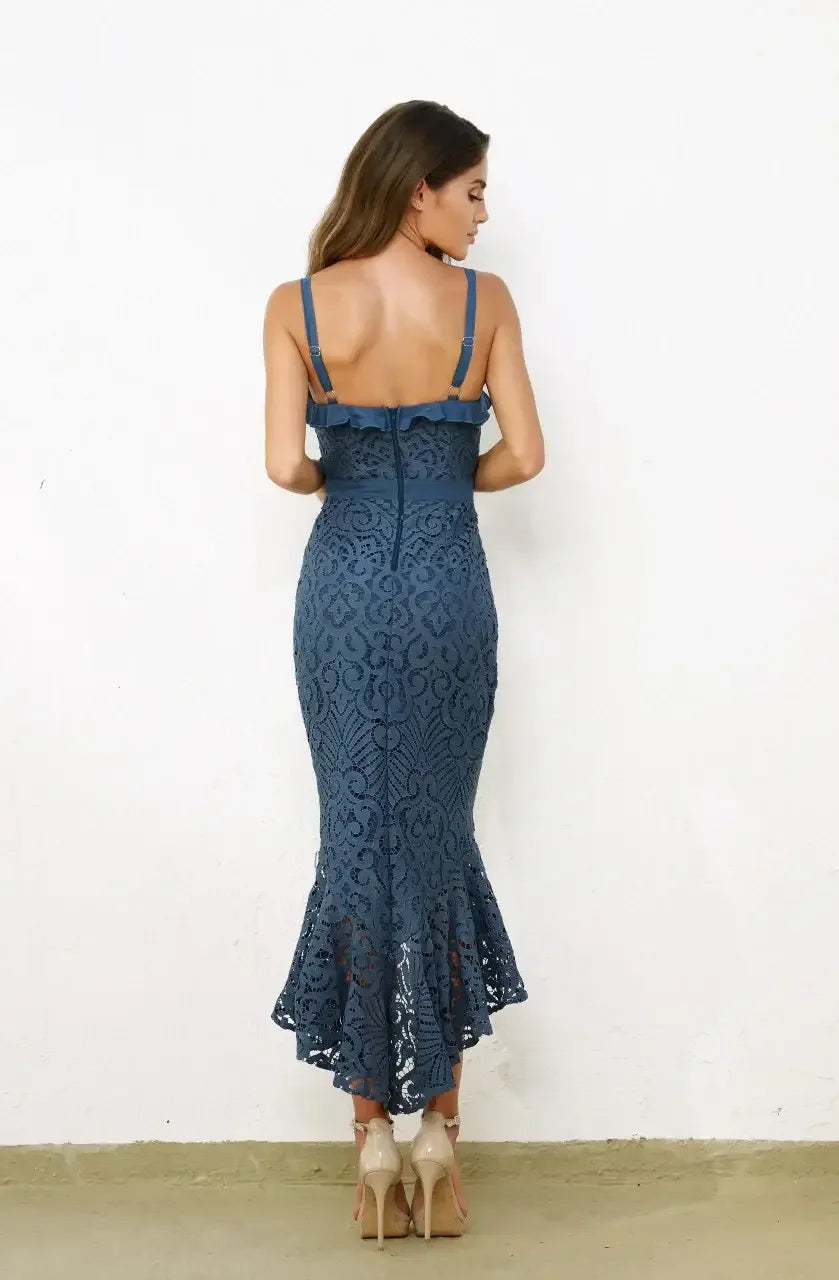 A woman in a fitted blue lace dress stands with her back to the viewer, showcasing the dress's ruffled straps and flared hem, against a plain white wall and light-colored floor.