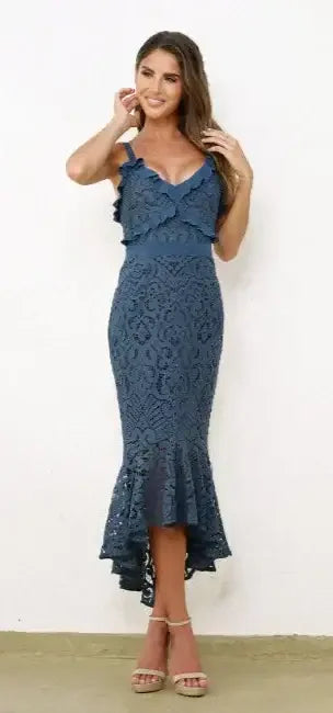 A woman in a dark blue lace dress with ruffles poses and smiles, gently touching her hair. She stands against a plain white wall and a light-colored floor.