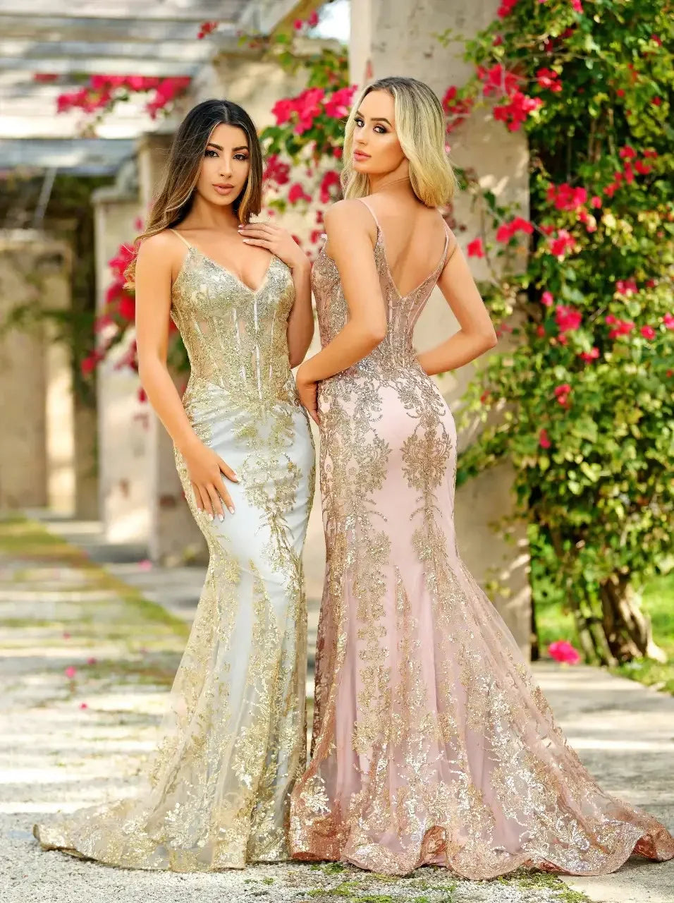 Two women are posing closely together, showcasing elegant, intricately designed evening gowns with golden embellishments. They stand in a picturesque garden, surrounded by blooming flowers and stone pathways.