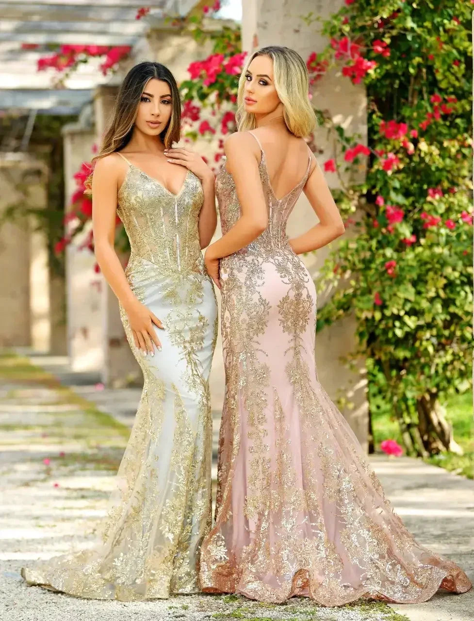 Two women pose in elegant, form-fitting evening gowns adorned with gold embellishments. They stand in a garden with vibrant pink flowers, creating a picturesque setting.