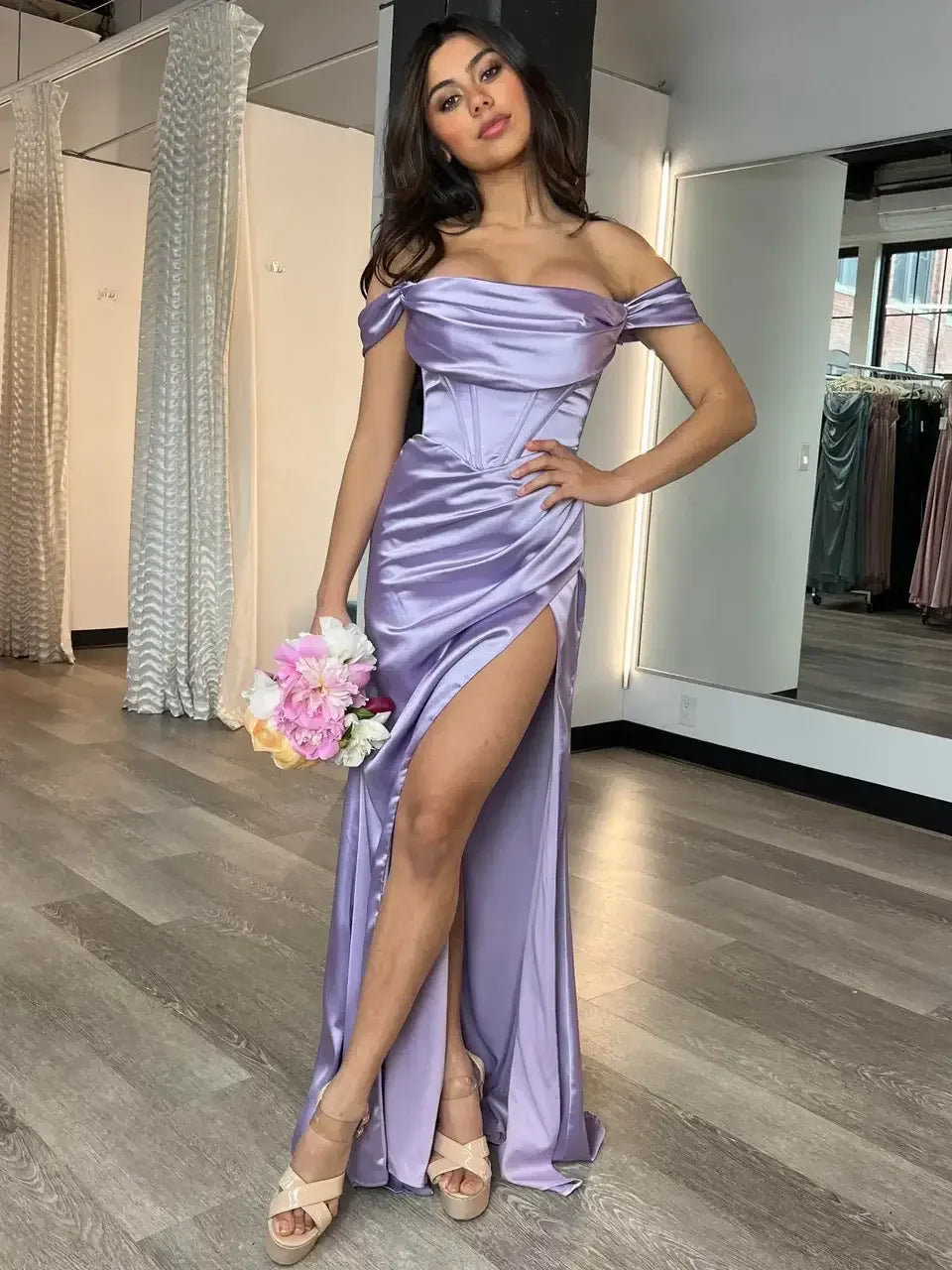 A woman is posing in a stylish, lavender, off-shoulder gown with a thigh-high slit, holding a bouquet of flowers, in a well-lit boutique with mirrors and draped fabrics.