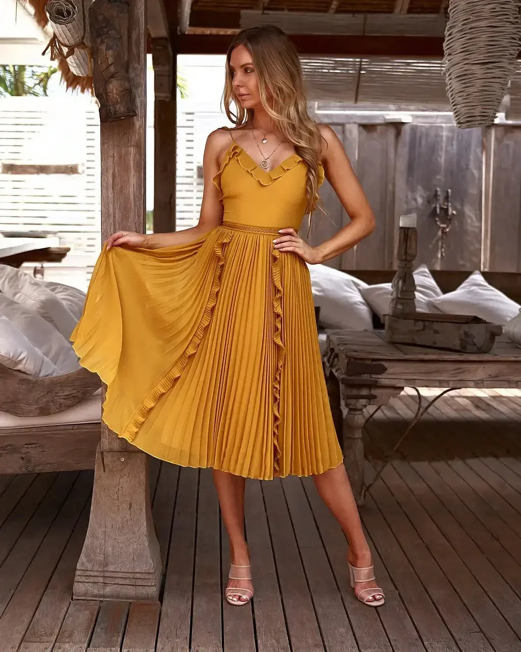 A woman in a flowing, yellow pleated dress stands gracefully, holding the hem. She is positioned in a stylish, rustic indoor setting with wooden accents and soft furnishings around her.