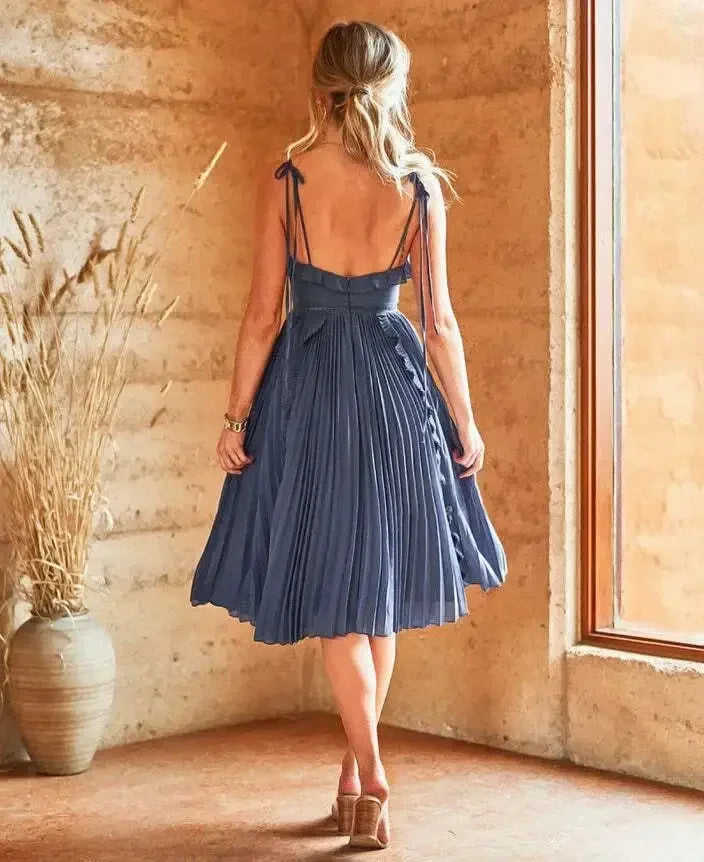 A woman in a pleated, blue dress stands with her back facing a window, showcasing the dress's flowing fabric. The environment features earthy tones and a nearby decorative vase.