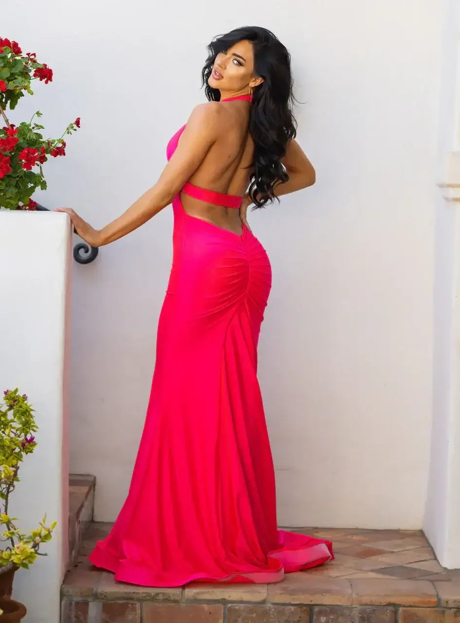 A woman is posing confidently while wearing a bright pink evening gown that highlights her curves. She stands against a white wall, with flowers nearby, creating a vibrant atmosphere.