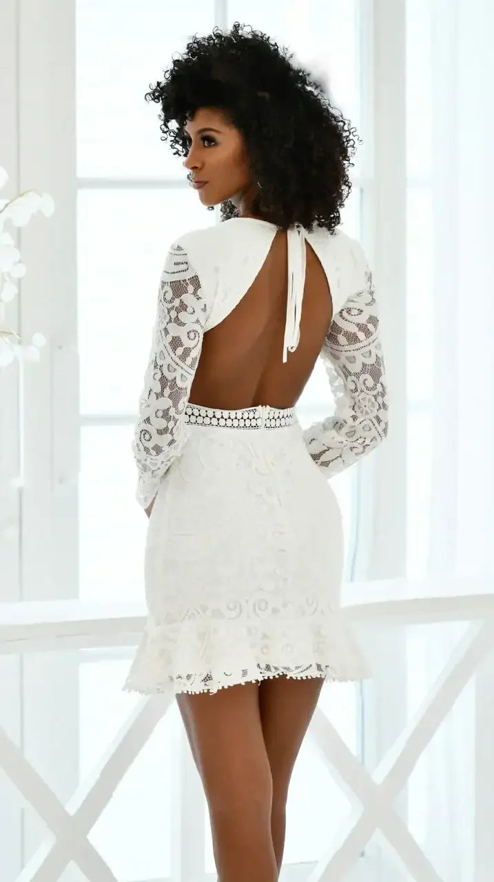 A woman stands with her back to the viewer, wearing a fitted white lace dress that features long sleeves and an open back. She is in a bright, airy room with large windows.