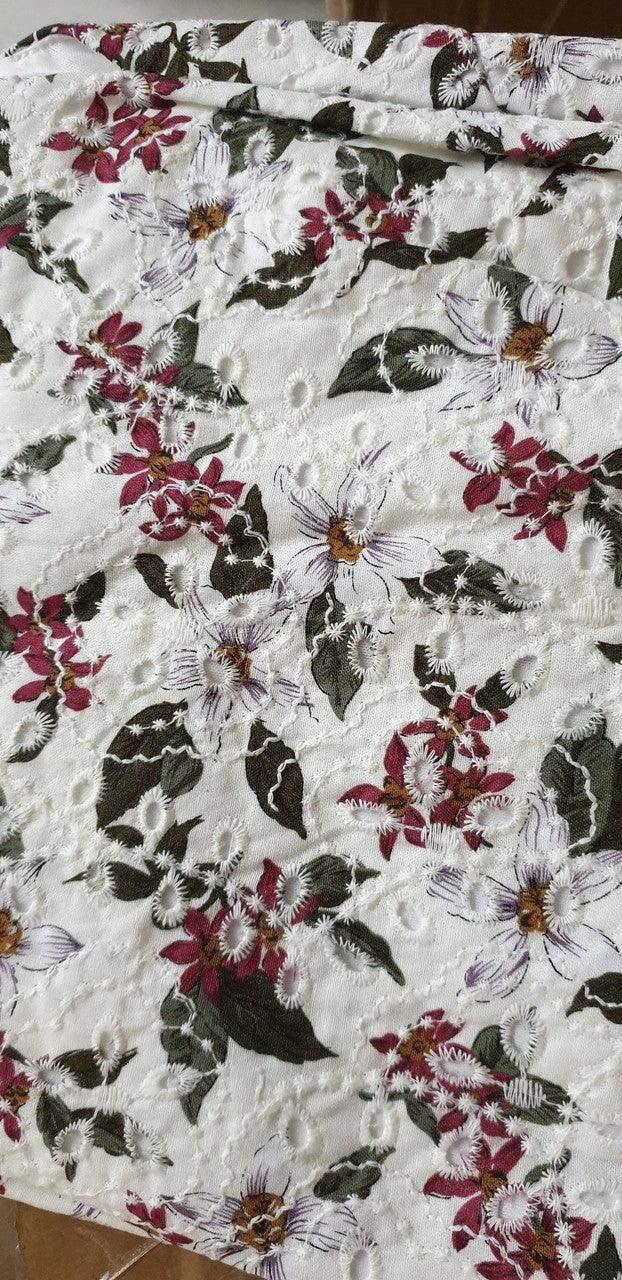 A floral fabric with intricate embroidery features vibrant flowers in shades of red, white, and green, draped across a surface, set against a neutral background.
