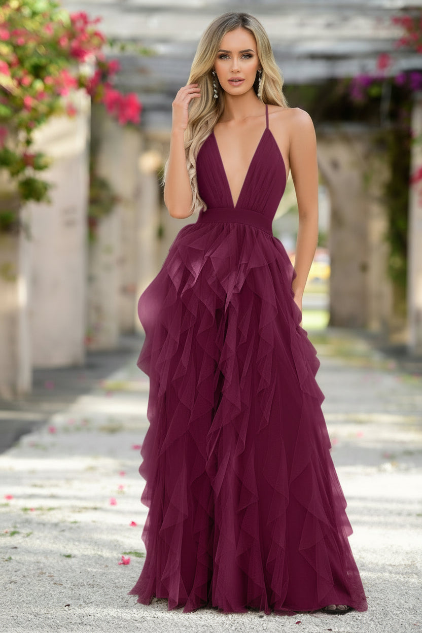 A woman wears a deep burgundy, floor-length dress with layered ruffles, standing confidently in a floral courtyard with soft lighting and greenery in the background, suggesting a romantic setting.