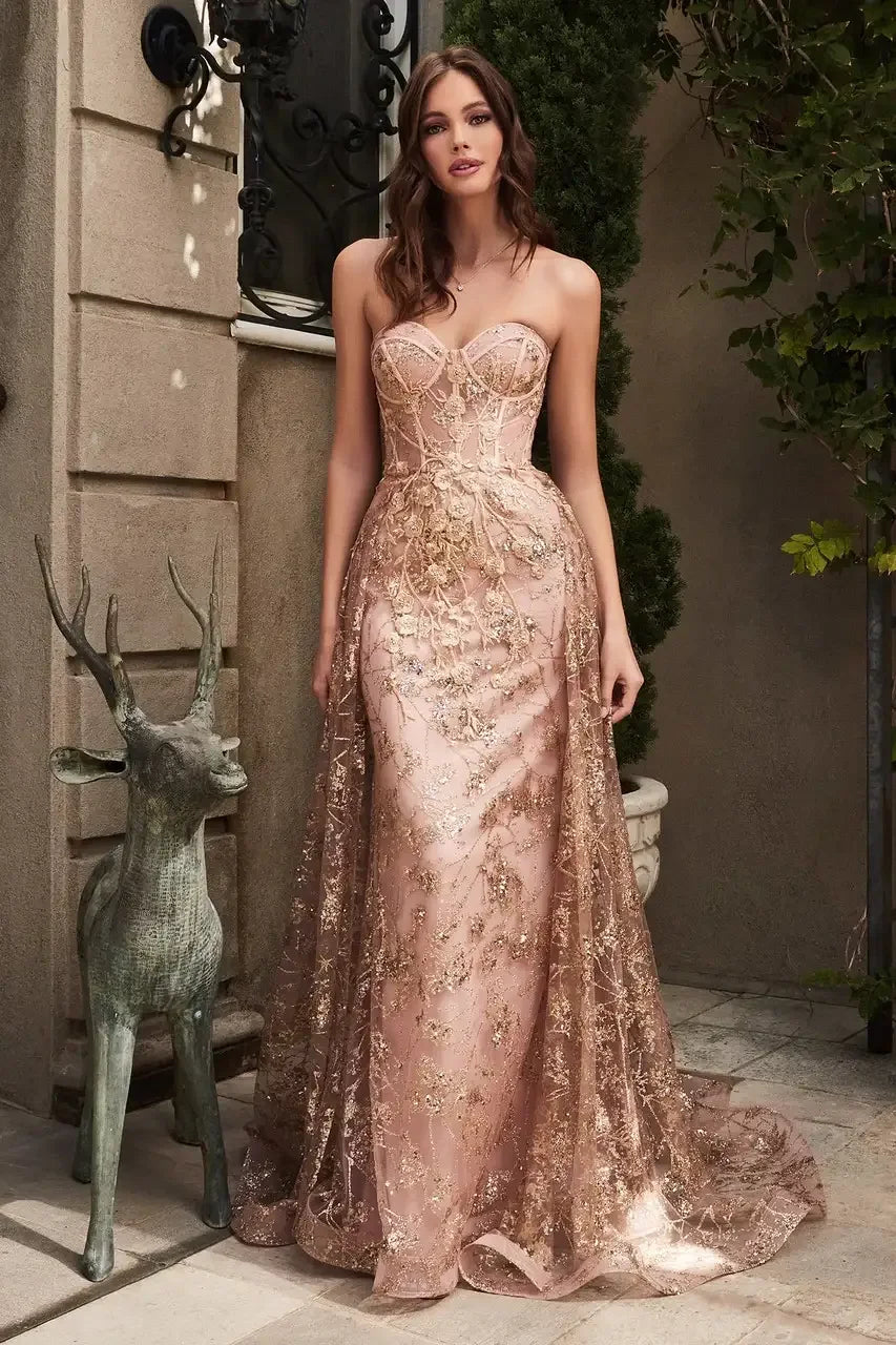 A woman stands elegantly in a pink, strapless gown adorned with gold embroidery. She poses beside a deer sculpture in a lush, garden-like setting.