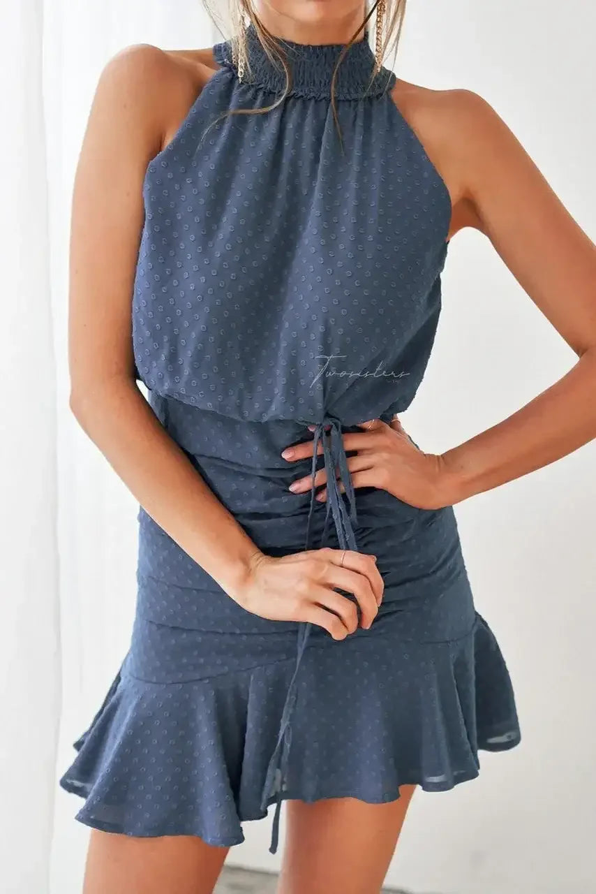 A person wears a sleeveless, dark blue dress with a ruffled hem, adjusting its drawstring while standing against a light backdrop, exuding a casual yet stylish vibe.