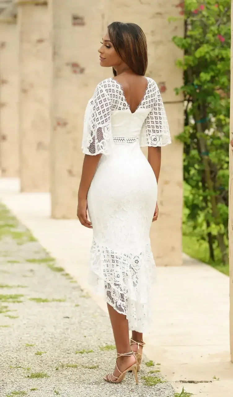 A woman in a white lace dress stands gracefully, facing away, on a stone path surrounded by greenery and textured stone columns, showcasing elegance and serenity in her pose.