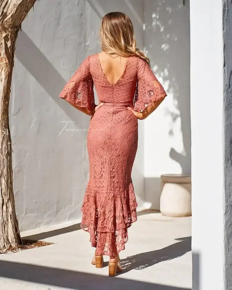 A woman stands wearing a long, pink lace dress with flared sleeves, showcasing a fitted silhouette and ruffled hem. The setting features a minimalist white wall and a light-filled space.