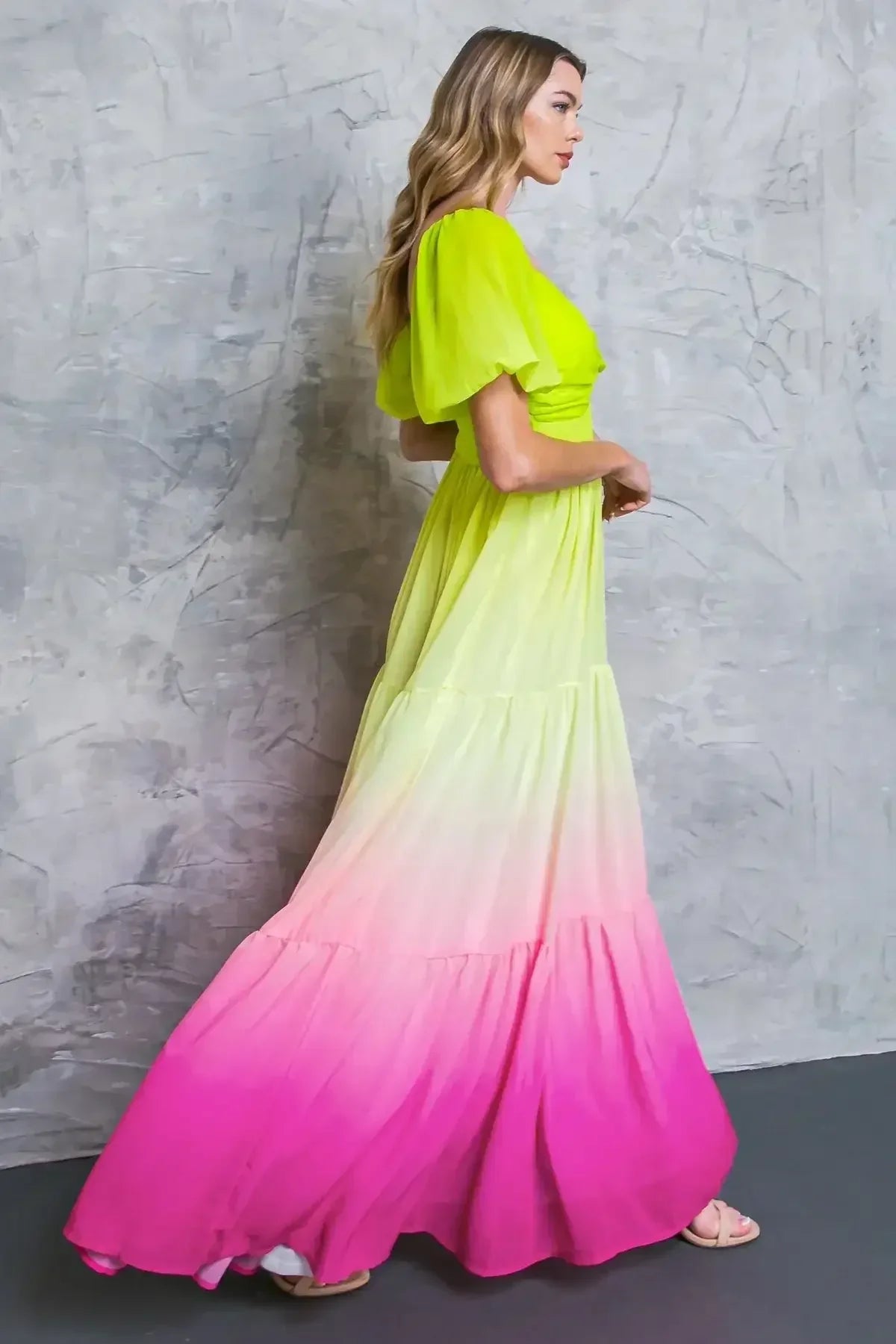 A woman in a vibrant, gradient dress transitions from bright yellow to pink as she walks against a textured, gray backdrop, showcasing the flow of the fabric and her elegant posture.